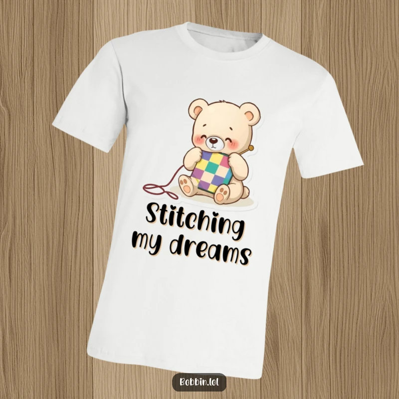Funny t-shirt print of a small bear happily stitching a colorful quilt square with a needle, showcasing gentle crafting energy.
